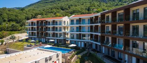Un hotel moderno de varios pisos, piscina en la azotea y montañas al fondo.