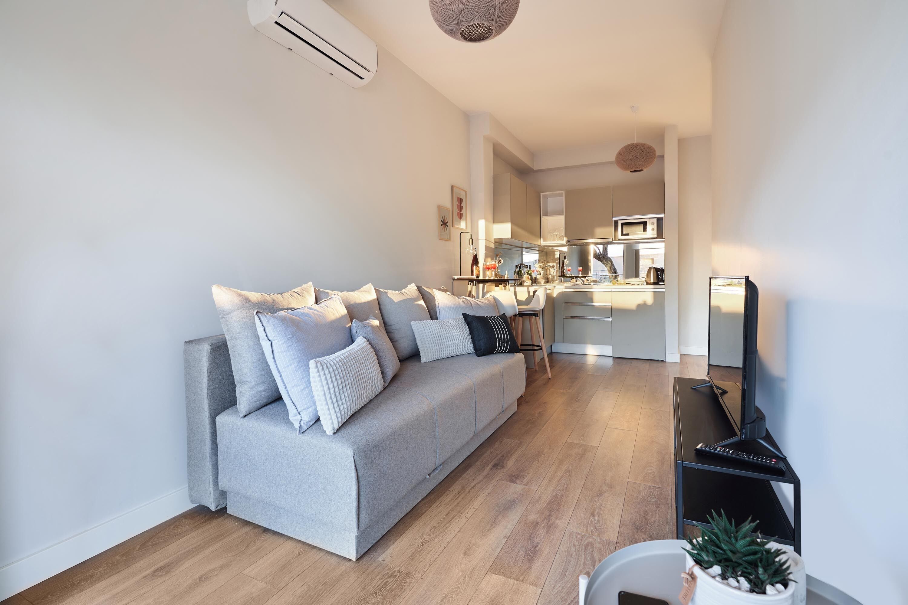 Una sala de estar moderna con un sofá gris, suelo de madera y una cocina al fondo.