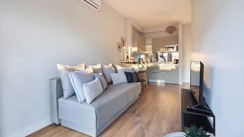 Una sala de estar moderna con un sofá gris, suelo de madera y una cocina al fondo.