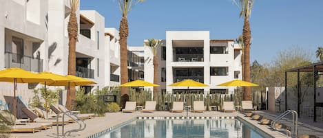 A pool area with lounge chairs, umbrellas, and a building with balconies.