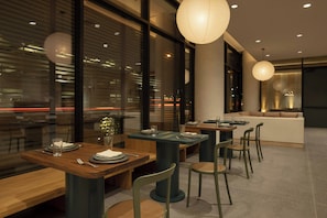 A modern dining area with tables and chairs, a sofa, and large windows. - Drift Palm Springs (PALM SPRINGS)