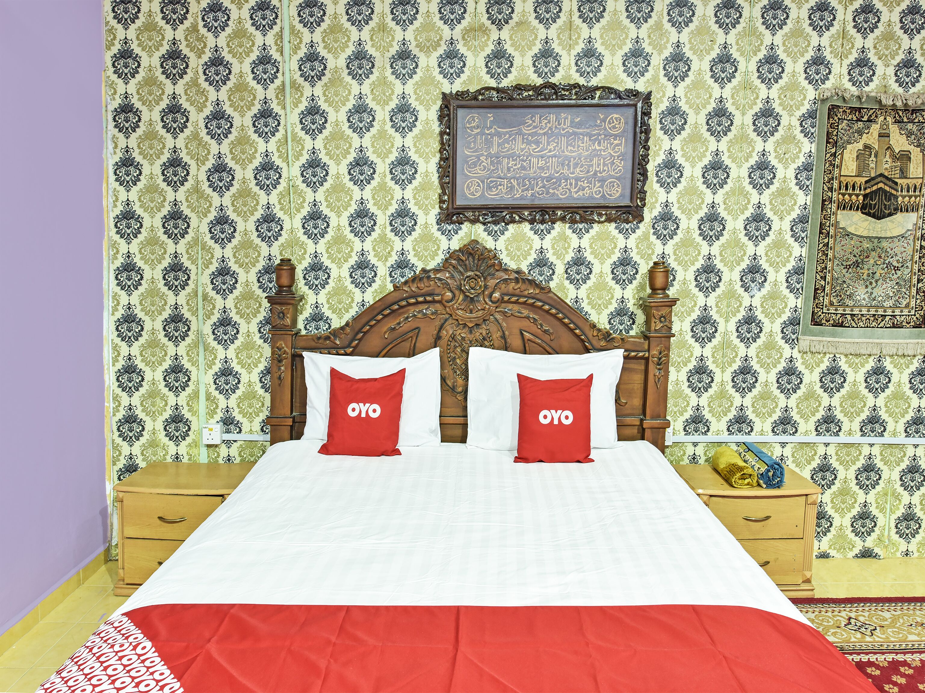 A neatly made bed with a red and white bedspread, two red pillows with the OYO logo, a wooden headboard, and patterned wallpaper.