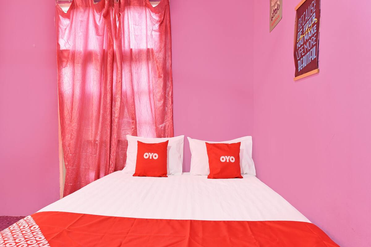 A bed with white and red bedding, two red pillows with the word "OYO" on them, and a pink wall with a poster.