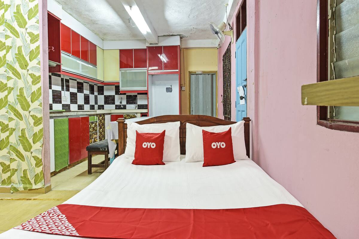 A room with a bed, a wooden headboard, and a red and white bedspread. There is a window with a wooden blind and a door with a blue frame.