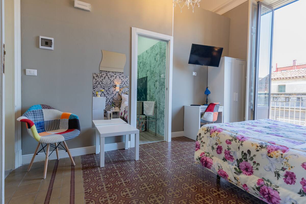A hotel room with a bed, a colorful chair, a small table, a TV, and a door leading to a bathroom.