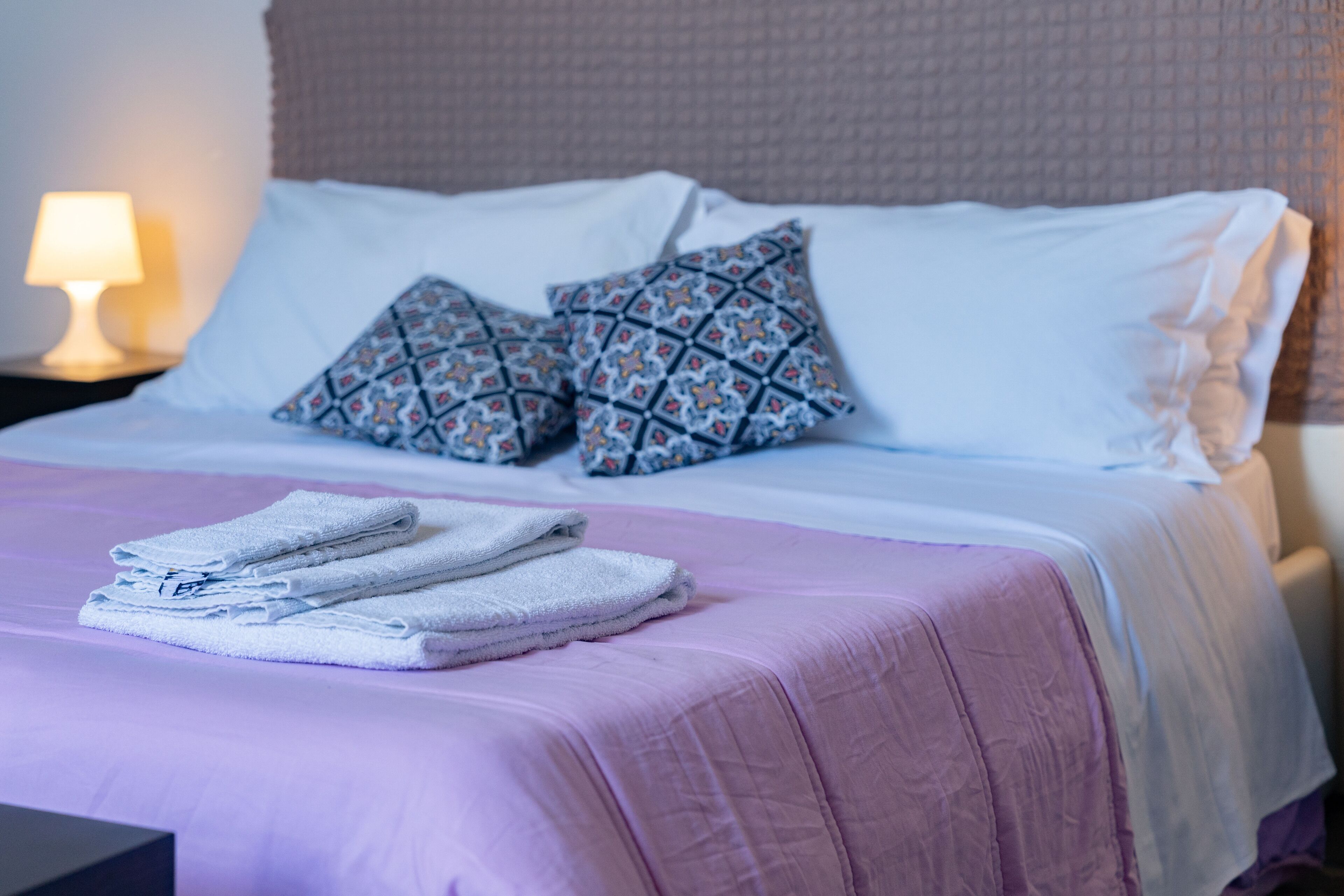 A neatly made bed with a pink bedspread and two patterned pillows, a stack of folded white towels, and a nightstand with a lamp.
