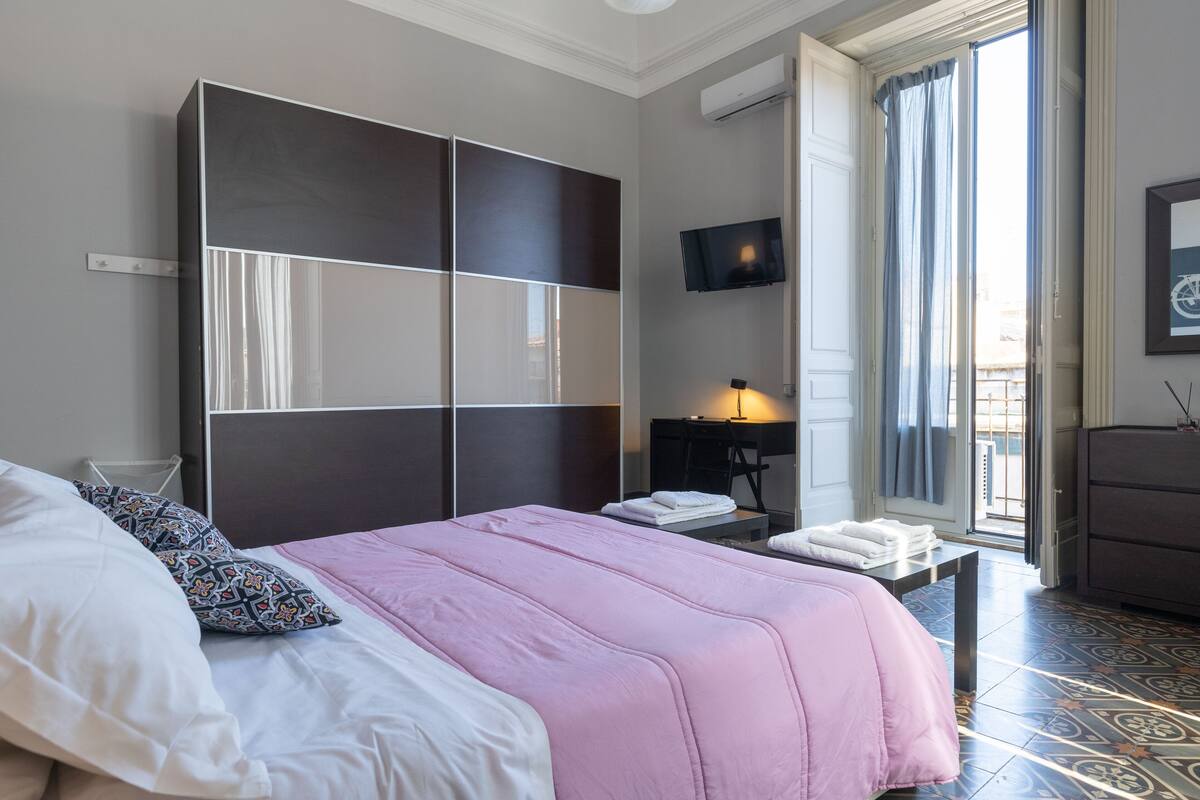 A modern hotel room with a large bed, a wardrobe, a desk, and a TV.