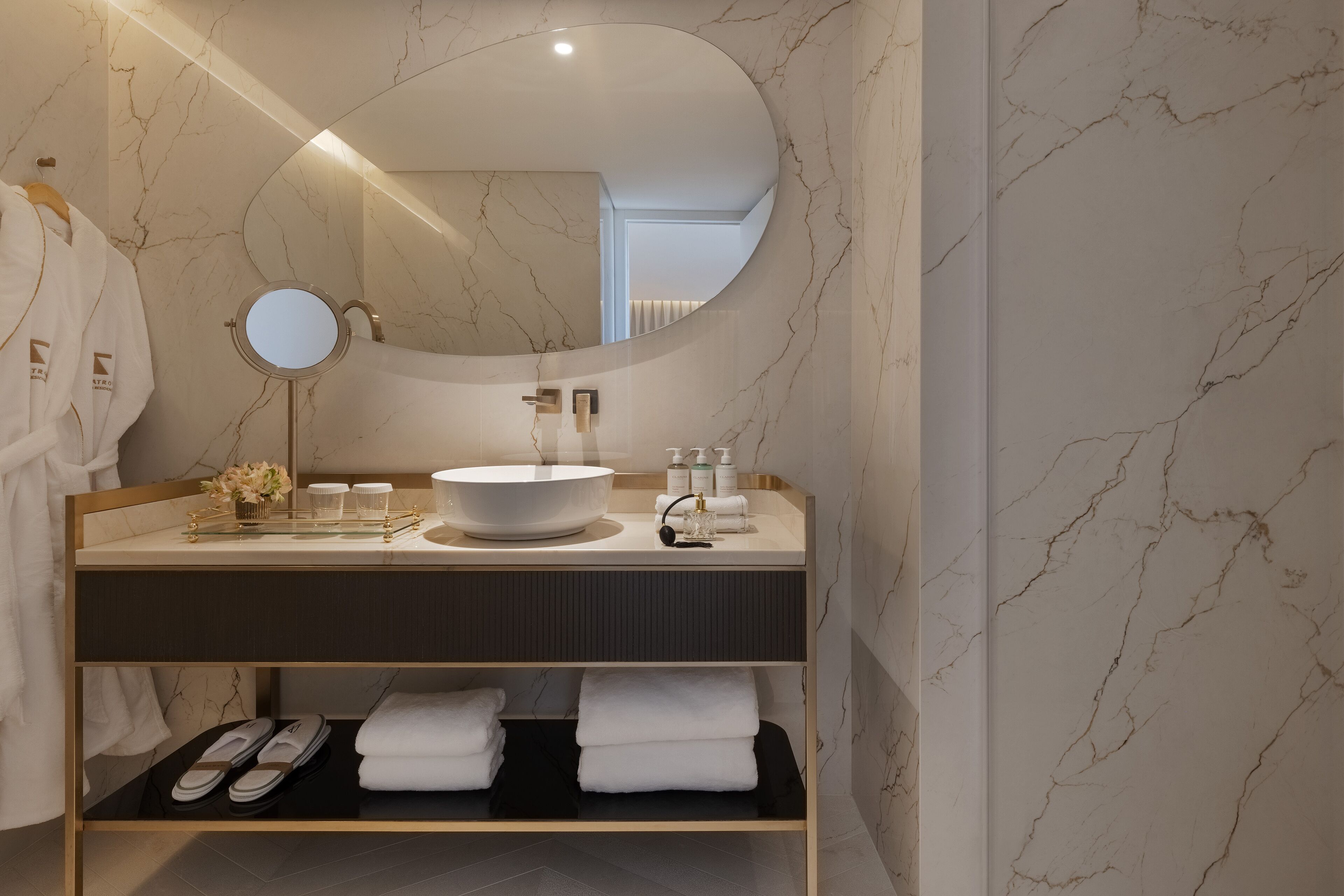 A modern bathroom with a marble backsplash, a round mirror, a large sink, and a shelf with towels and slippers.