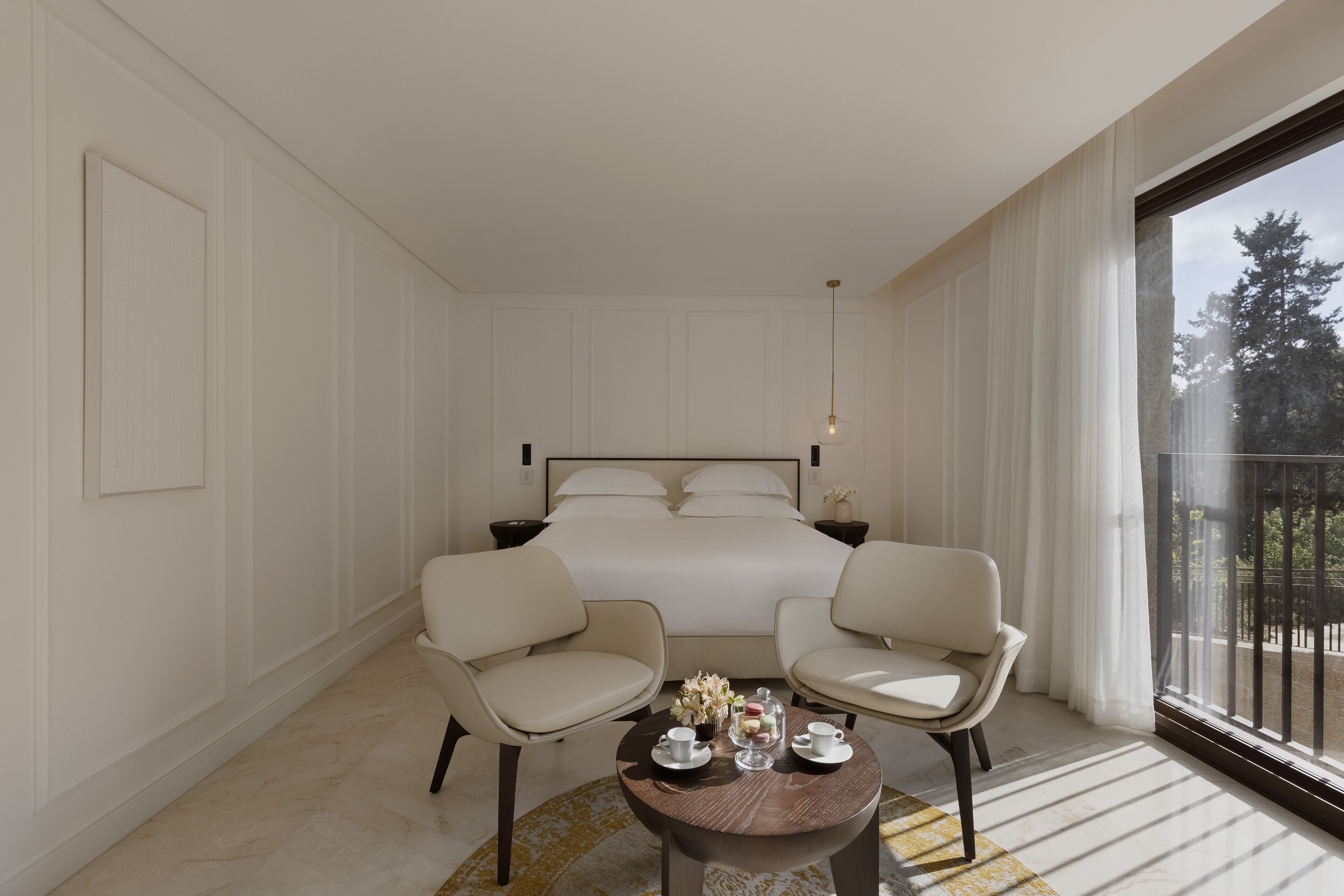 Una habitación de hotel con una cama grande, dos sillones, una mesita con un juego de té y vistas a árboles por la ventana.