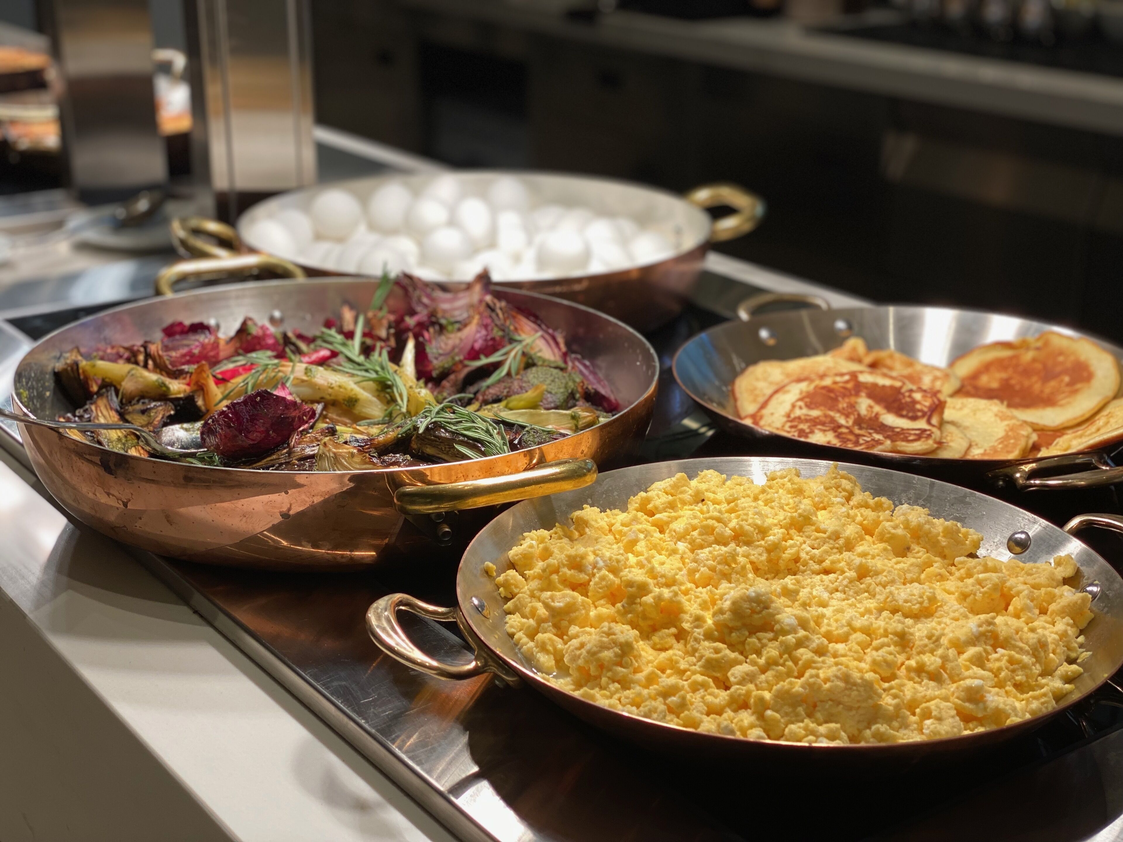 A buffet with various dishes including scrambled eggs, roasted vegetables, and pancakes.