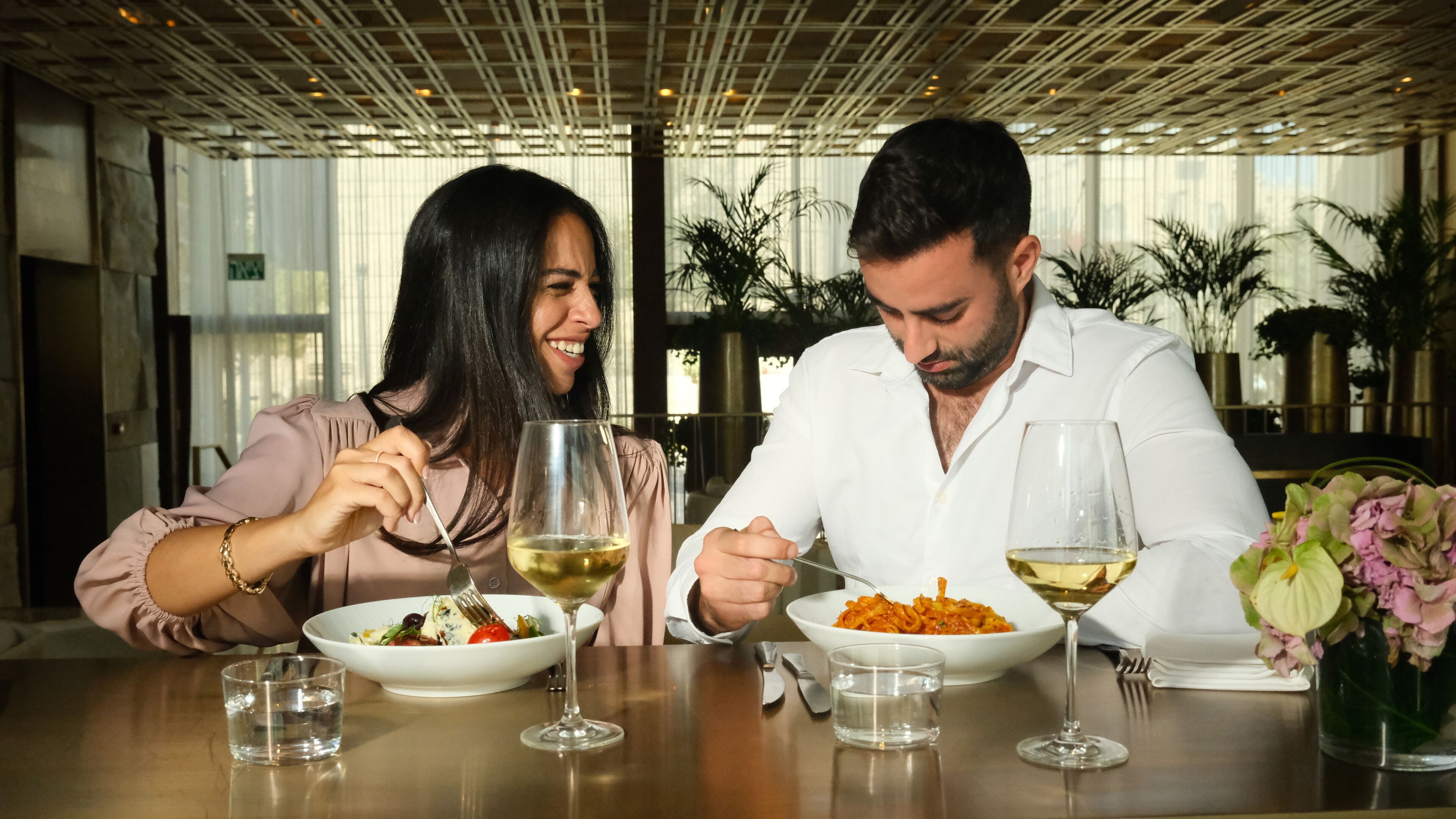 Una mesa de comedor con dos personas, un bol de ensalada, un plato de pasta y dos copas de vino.