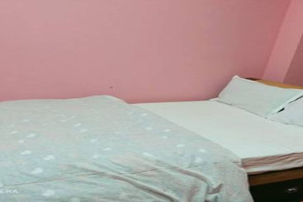 Un lit simple avec un matelas blanc et une couette de couleur claire, dans une pièce aux murs roses.
