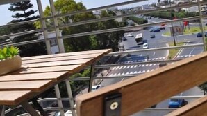Un balcon avec une table et des chaises en bois, donnant sur un parking et une route où circulent des voitures.
