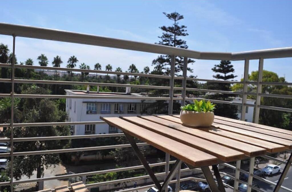 A balcony with a wooden table and a potted plant, overlooking a street with cars and buildings.