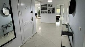 Un couloir avec un sol carrelé, une porte, un miroir et un luminaire suspendu.