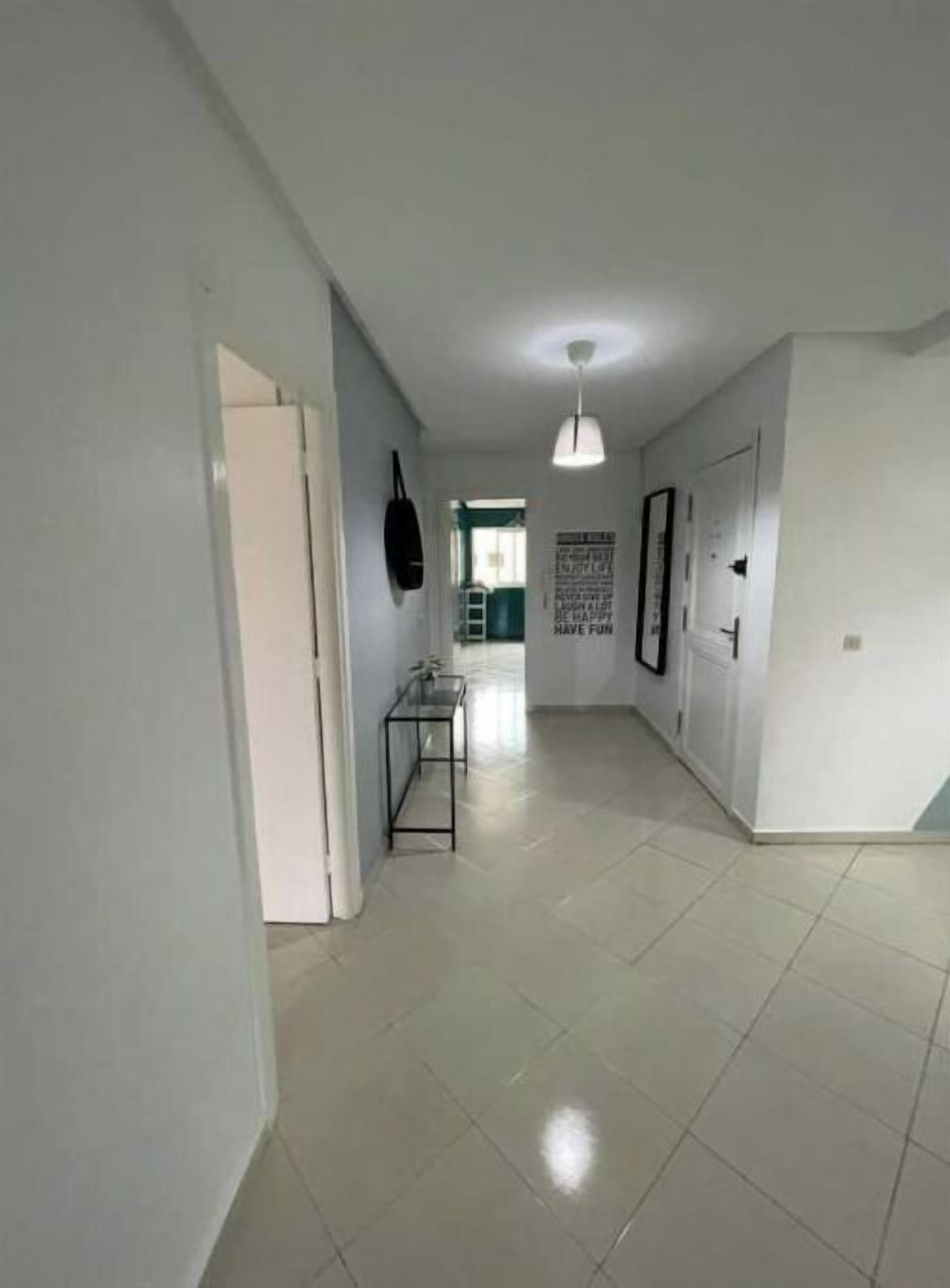 A hallway with a tiled floor, a door on the left, a small table with a chair, and a hanging bag on the wall.