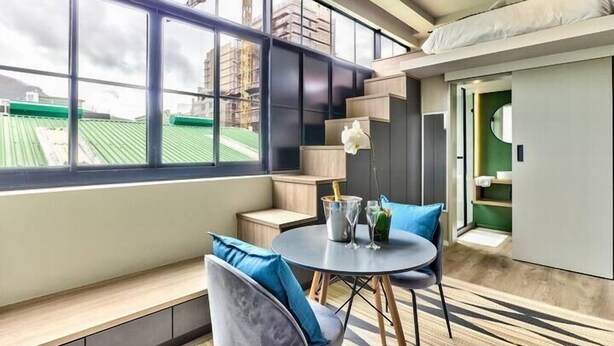 A modern hotel room with a large window, a dining area with a round table, and a built-in kitchen. - The Harri 209 (Cape Town)