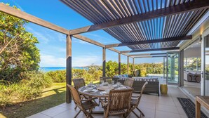 An outdoor patio with a round table set for dining, surrounded by wicker chairs, under a wooden pergola with a view of the sea.