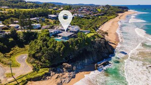 An aerial view of a coastal property with a large house surrounded by lush greenery, a beach, and a clear view of the ocean.