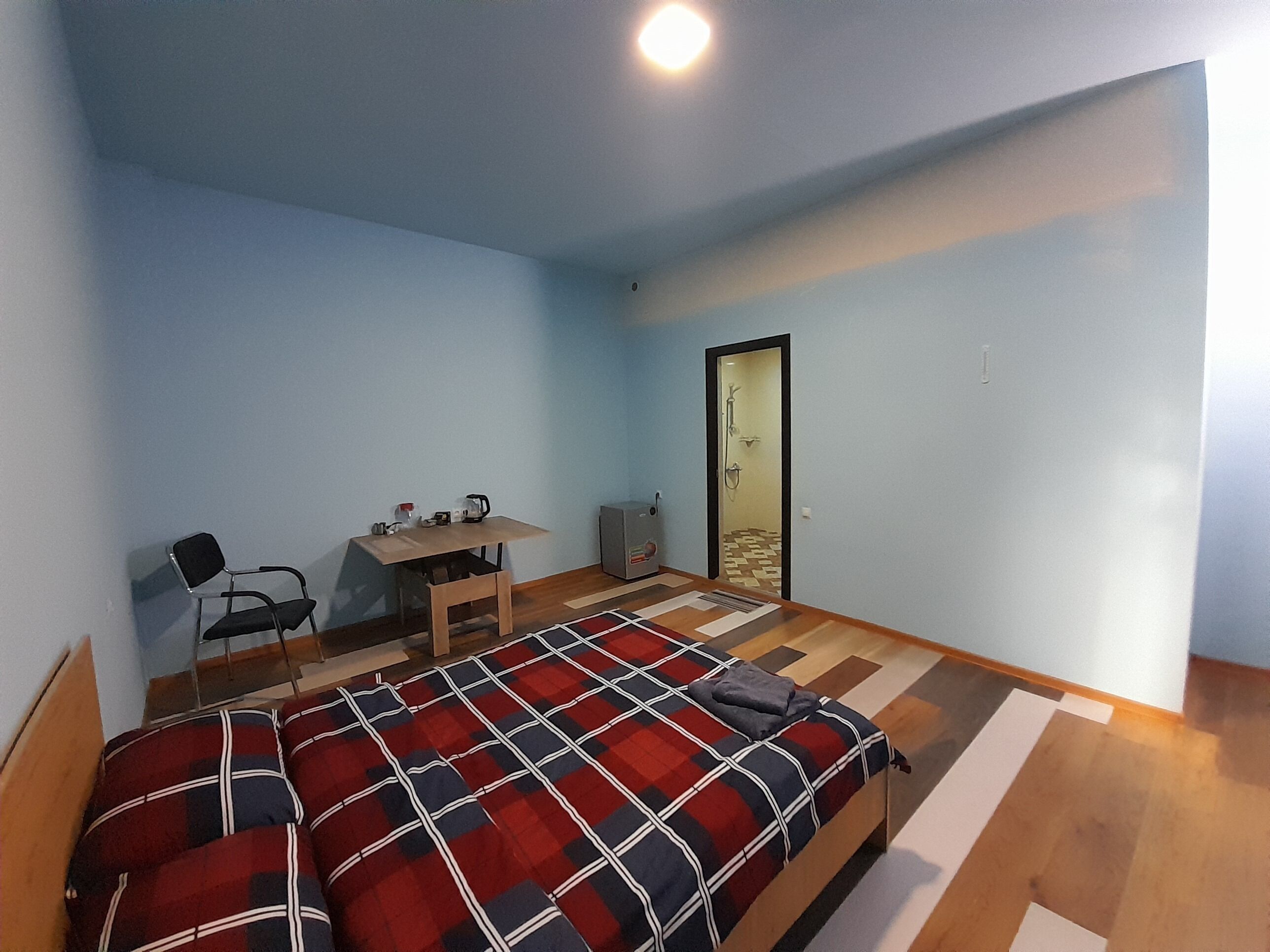 A small, well-lit room with a bed, a desk, a chair, and a shower area.
