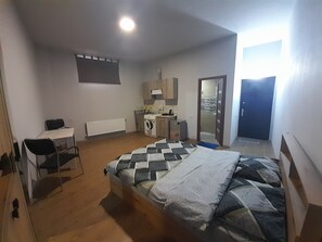 A small, well-lit room with a bed, a desk, a chair, a washing machine, and a small kitchenette.