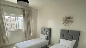 Une chambre moderne avec deux lits, une grande fenêtre avec des rideaux, une horloge murale ronde et un luminaire suspendu.