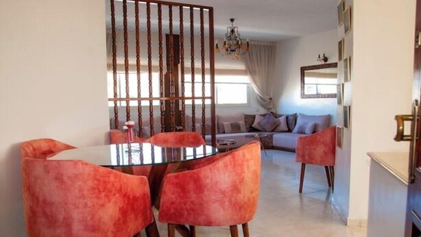Un espace repas avec une table ronde et six chaises, un ensemble de canapés à l’arrière-plan et un coin cuisine sur la droite.