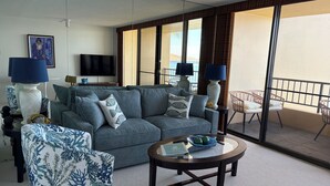 Ein modernes Wohnzimmer mit einem grauen Sofa, einem gemusterten Sessel und einem runden Couchtisch. Zwei Lampen, ein Flachbildfernseher und ein großes Fenster mit Vorhängen sind ebenfalls vorhanden.