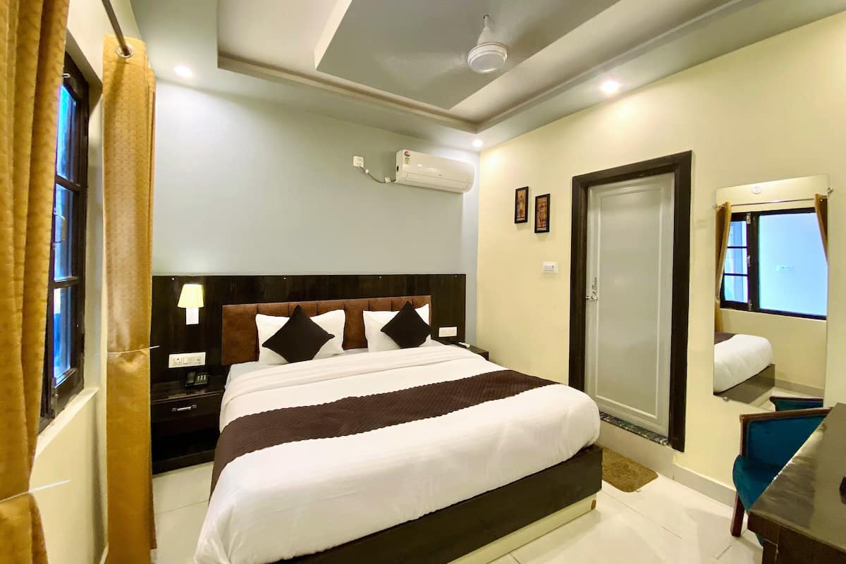 A hotel room with a bed, bedside table, lamp, air conditioning unit, and a window with curtains.