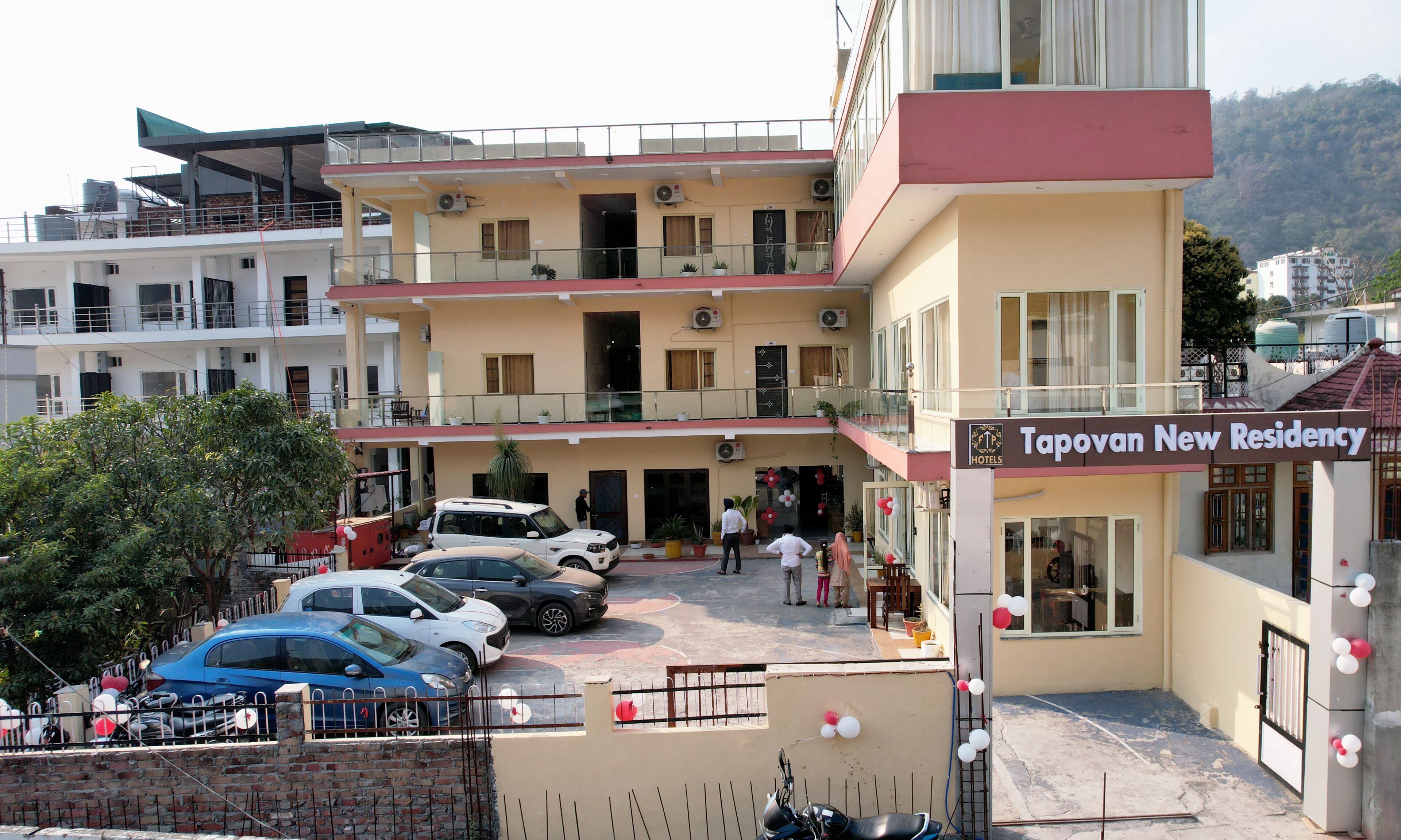 A multi-story hotel with a sign reading "Tapovan New Residency." There are several cars parked in front, and the building has multiple balconies with railings.