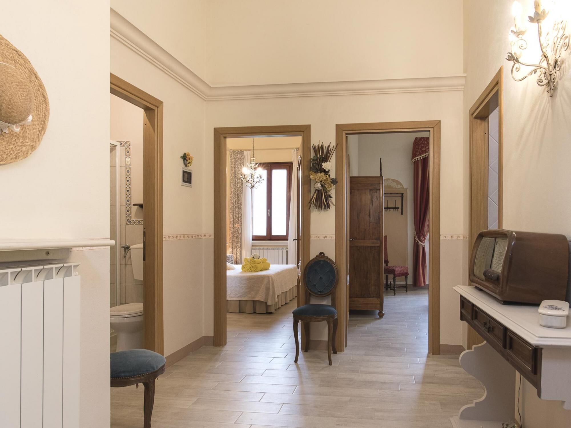 A hallway with a radiator, a bed with a white bedspread, a chair, a wooden wardrobe, and a television on a stand.