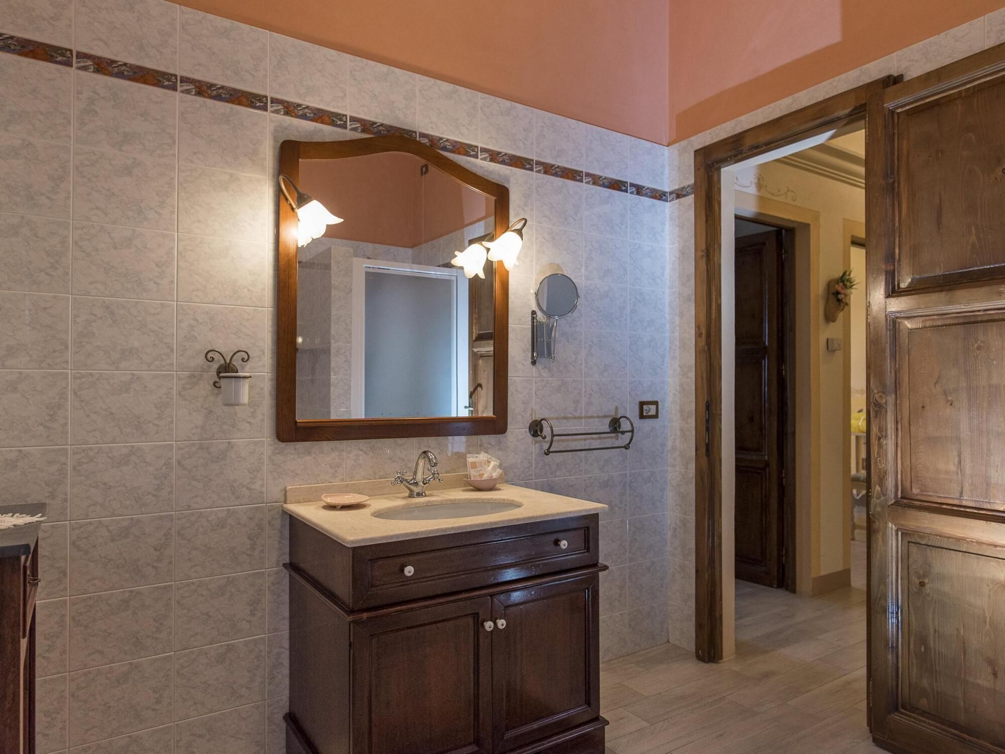 A bathroom with a vanity, sink, and mirror. There is a towel rack and a small round mirror on the wall. The floor is tiled, and the walls are covered with large tiles.