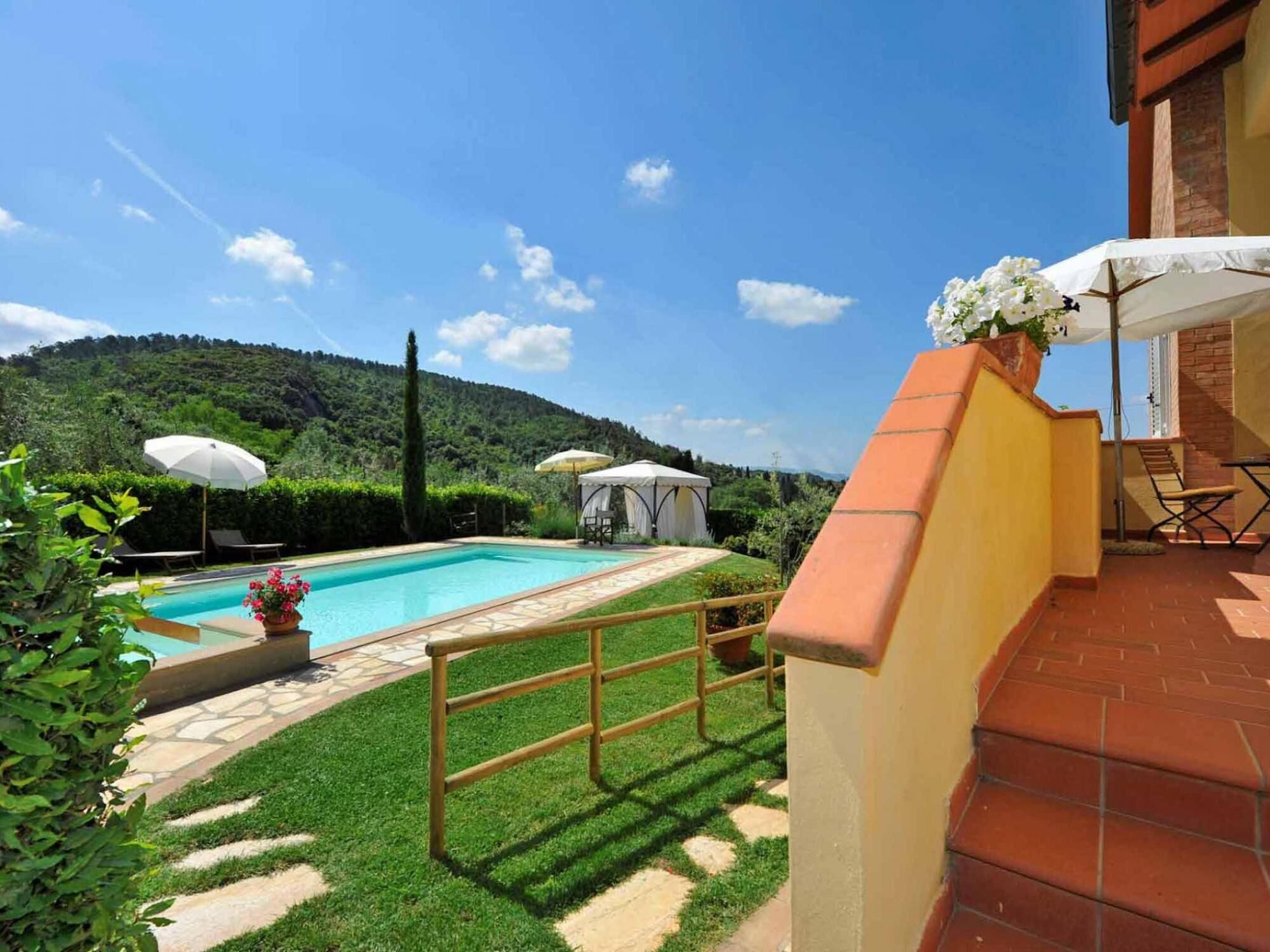 Ein Swimmingpool, umgeben von einer gepflegten Rasenfläche, einer überdachten Sitzgruppe und Blick auf eine hügelige Landschaft.