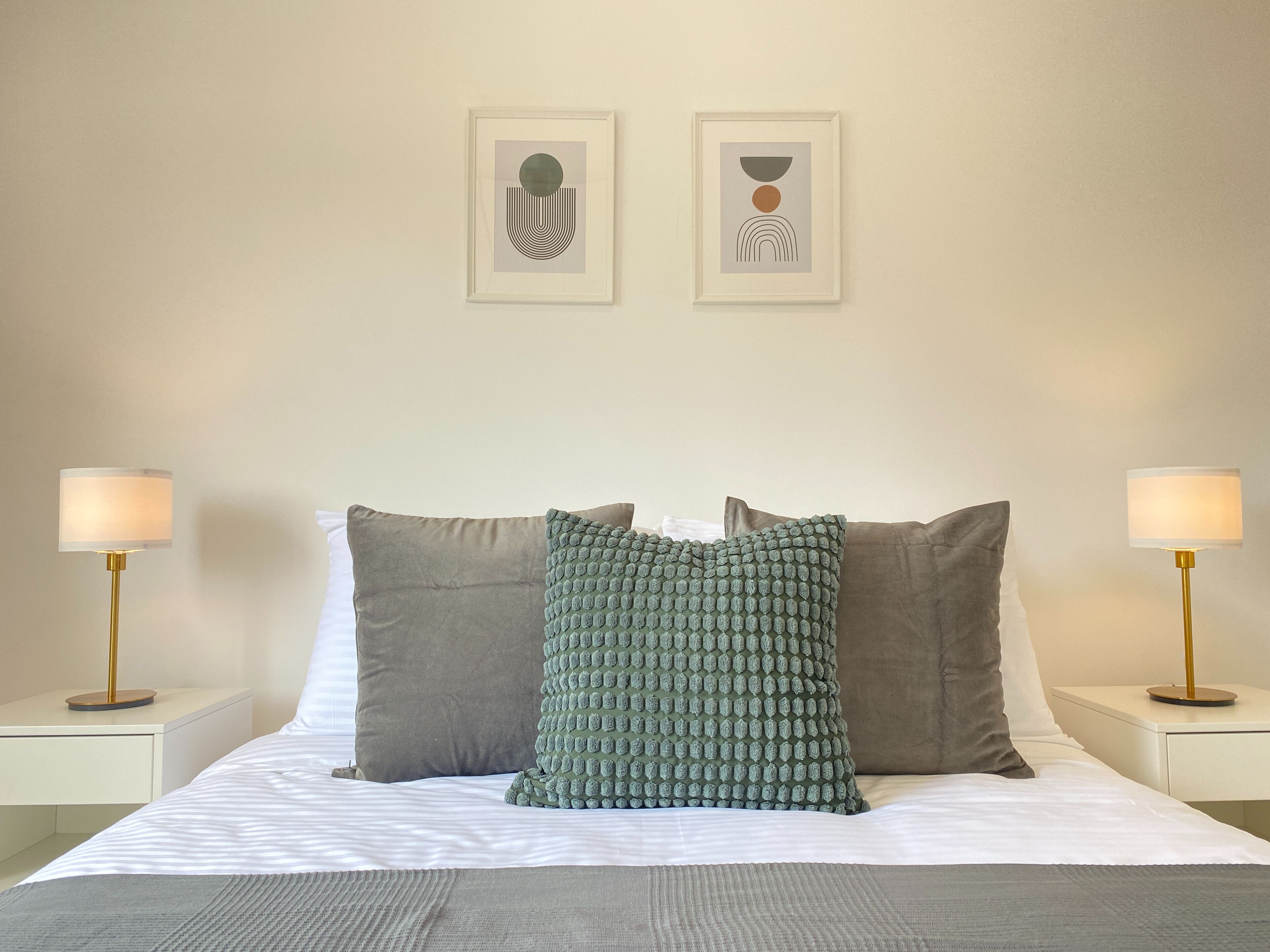 A neatly made bed with two pillows, two bedside lamps, and two framed abstract artworks on the wall.