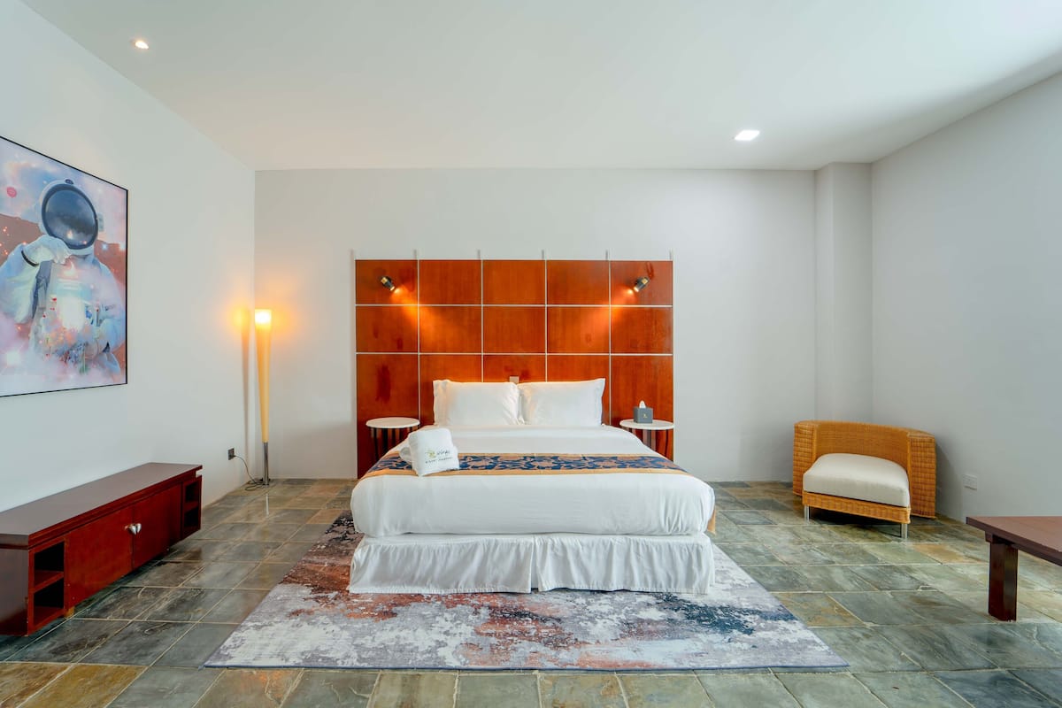 Una habitación de hotel moderna con una cama grande, un cabecero de madera, una mesita consola roja, una silla de mimbre y una pequeña mesa.