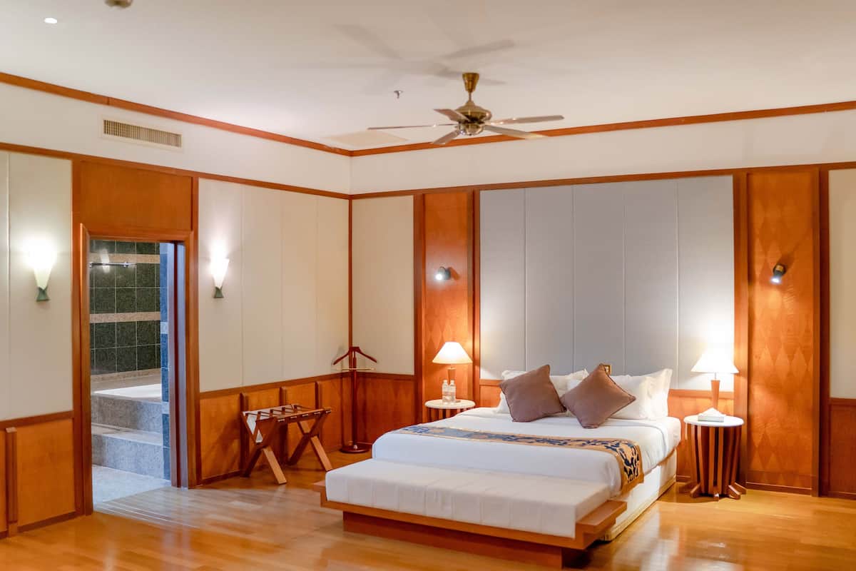 Habitación de hotel con una cama grande, dos mesitas de noche con lámparas, un ventilador de techo y vistas a un pasillo con una puerta que da al exterior.