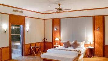 Habitación de hotel con una cama grande, dos mesitas de noche con lámparas, un ventilador de techo y vistas a un pasillo con una puerta que da al exterior.