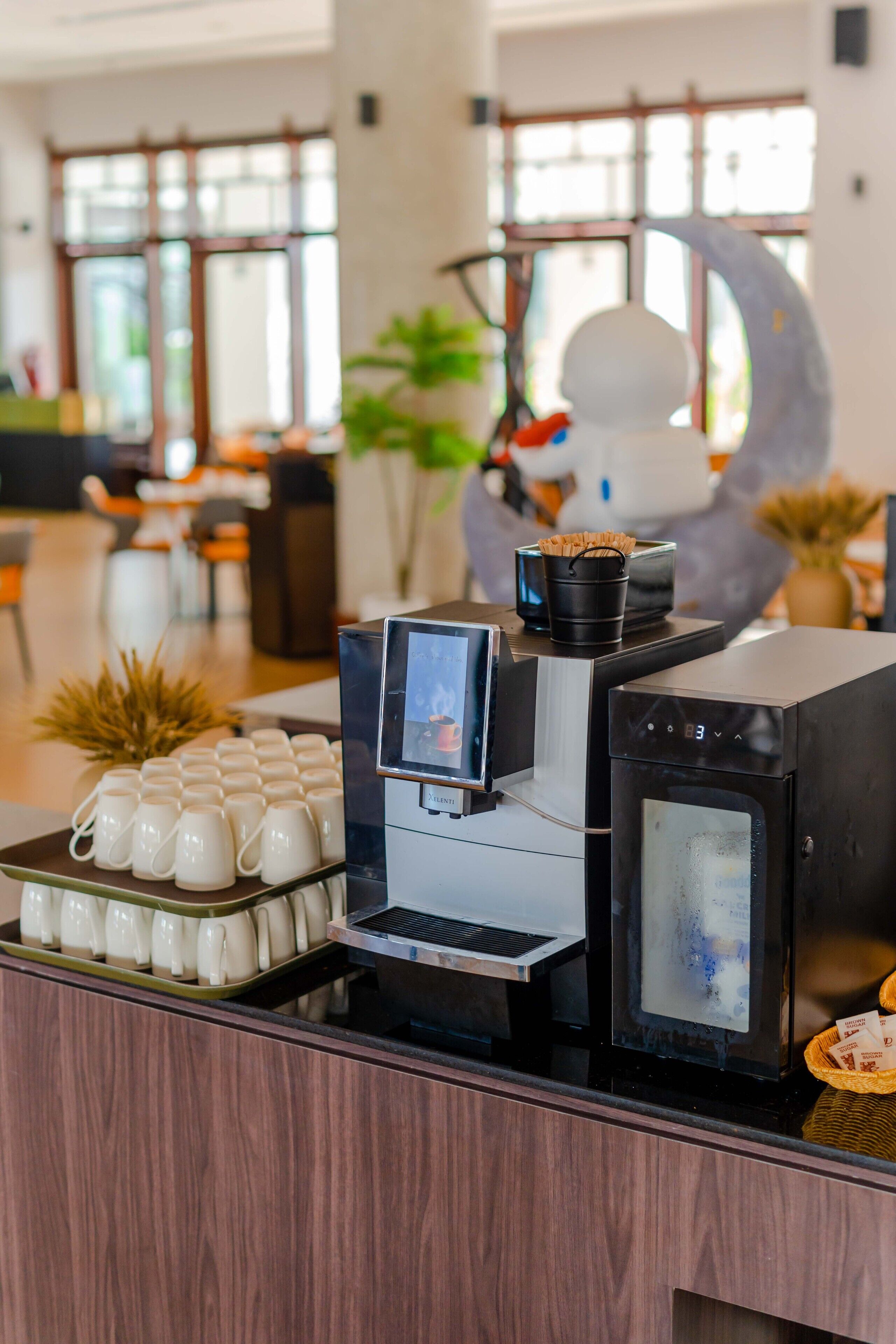 Uma estação de café com uma máquina de café, uma tela digital e uma bandeja de xícaras brancas.