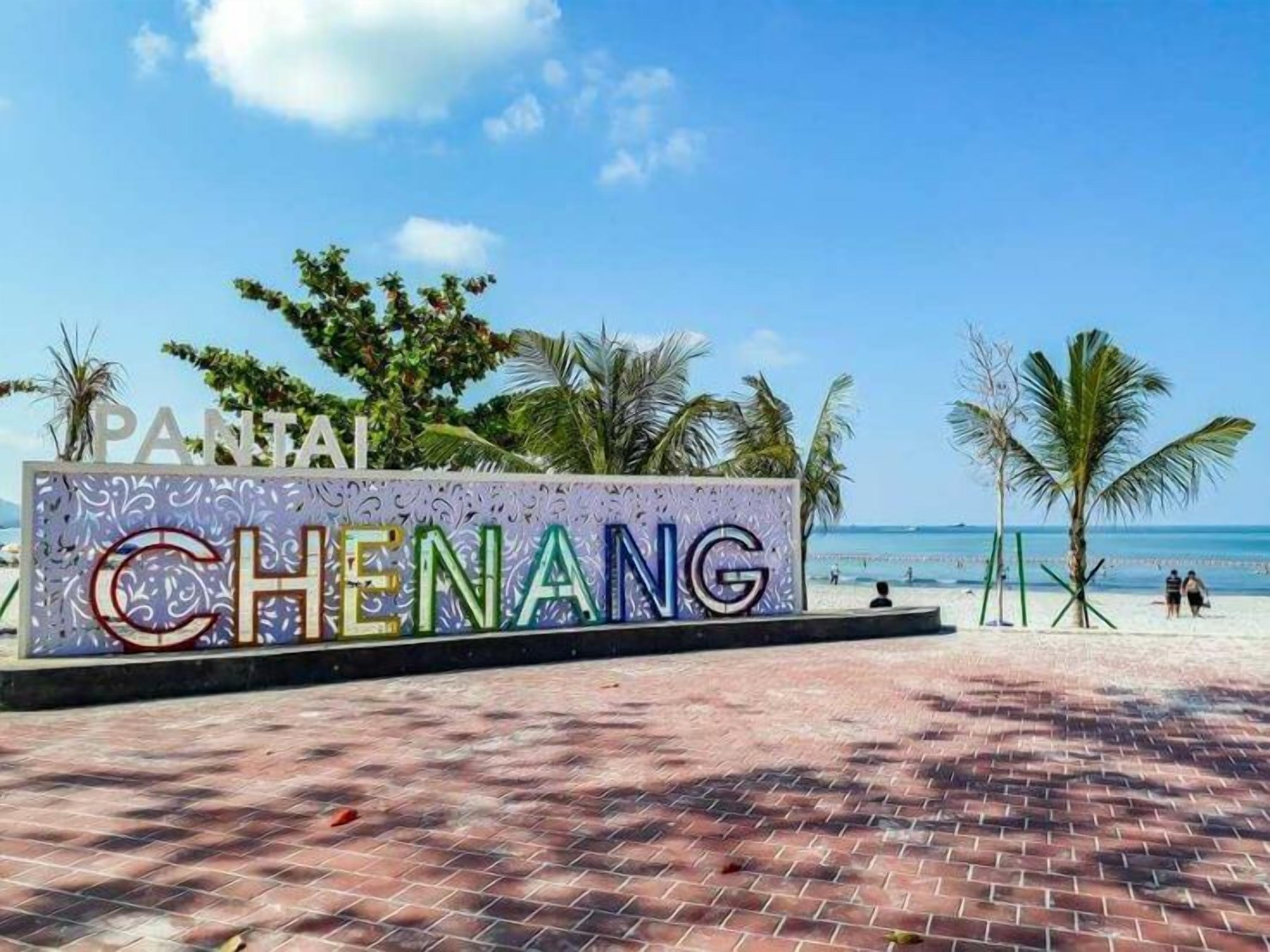Et skilt ved stranden med teksten "PANTAI CHENANG", omgivet af palmer og en klar blå himmel.