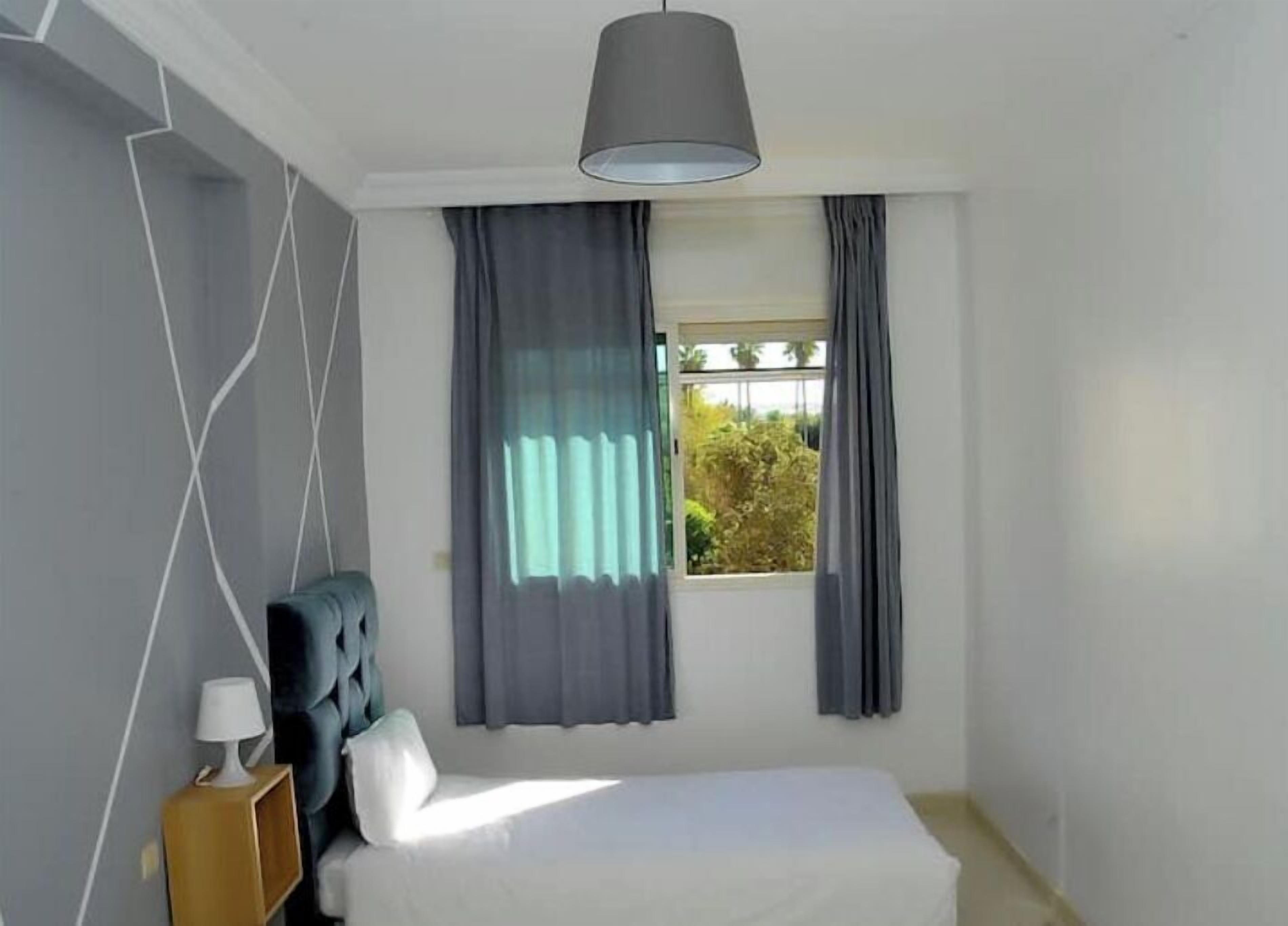 A bedroom with a bed, a bedside table with a lamp, a hanging light fixture, and a window with curtains.