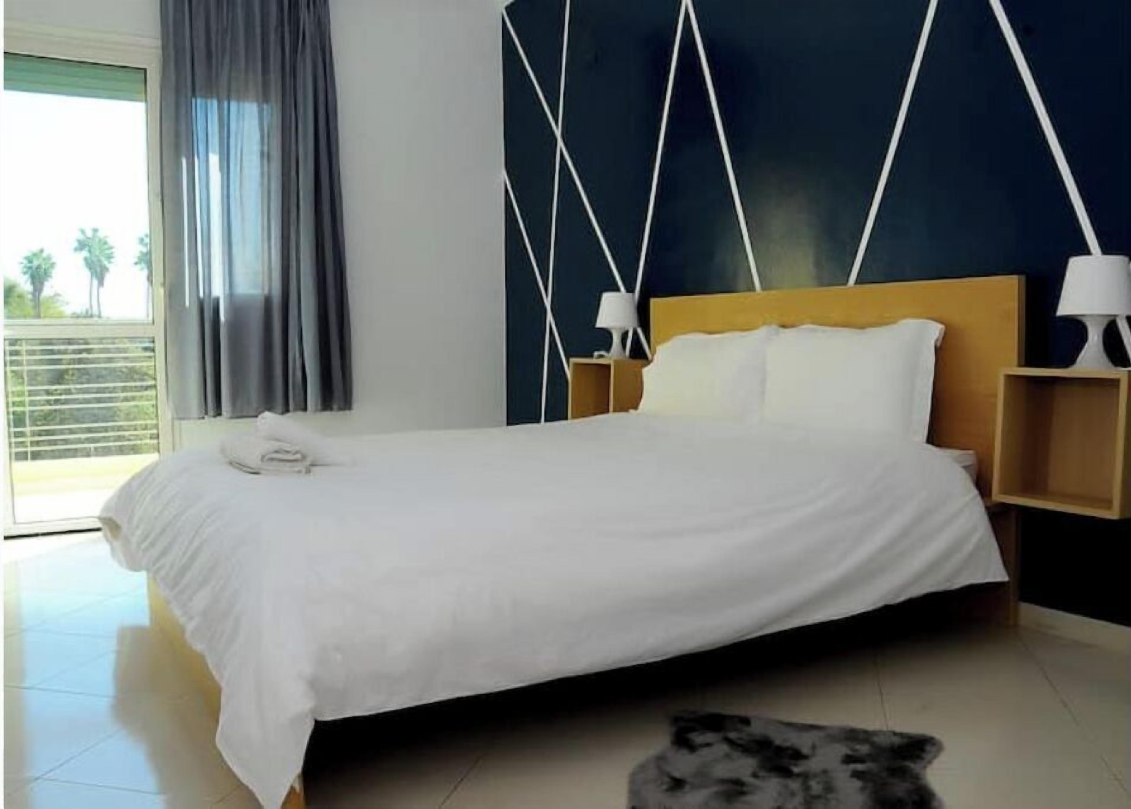 A hotel room with a large bed, two bedside tables with lamps, a view of palm trees through the window, and a dark wall with a geometric pattern.