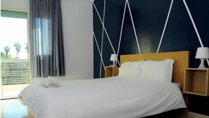 Et hotelværelse med en stor seng, to sengeborde med lamper, udsigt til palmer gennem vinduet og en mørk væg med et geometrisk mønster.