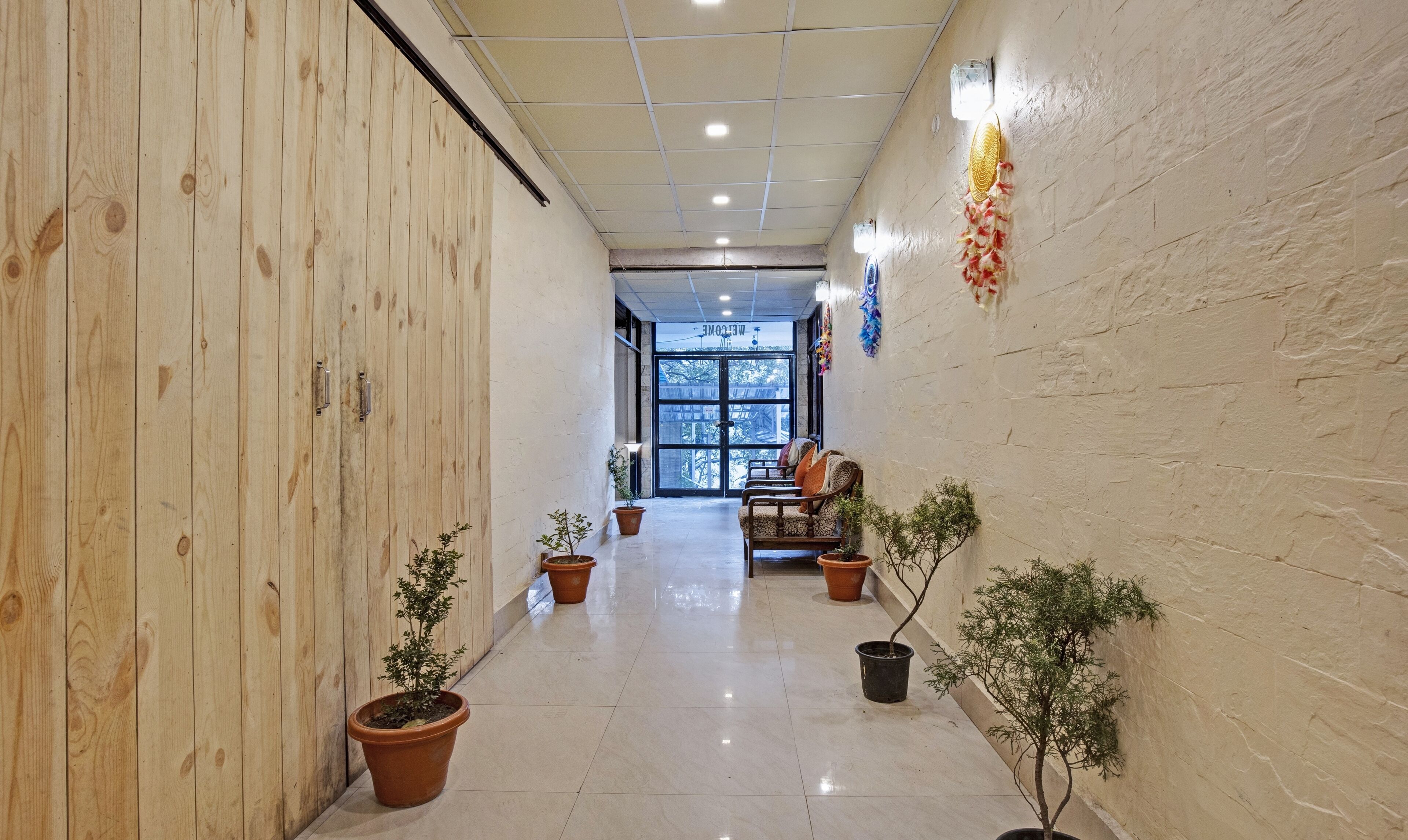 A corridor with wooden paneling, potted plants, a seating area with chairs, and a view of the outdoors through a glass door.