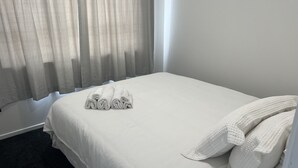 Una cama bien hecha con sábanas y almohadas blancas, un suelo alfombrado oscuro y cortinas translúcidas entreabiertas.