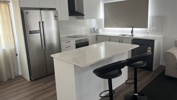 Una cocina moderna con armarios blancos, un refrigerador de acero inoxidable, un horno empotrado y una campana extractora negra. Hay una isla grande con un taburete negro.