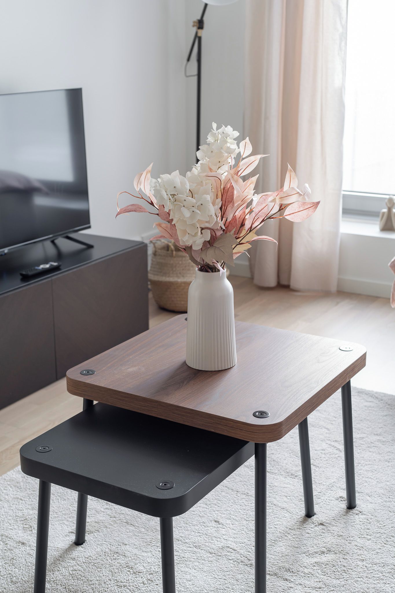 Una sala moderna con televisor, un jarrón blanco con flores, una mesa de centro de madera y un otoman negro.