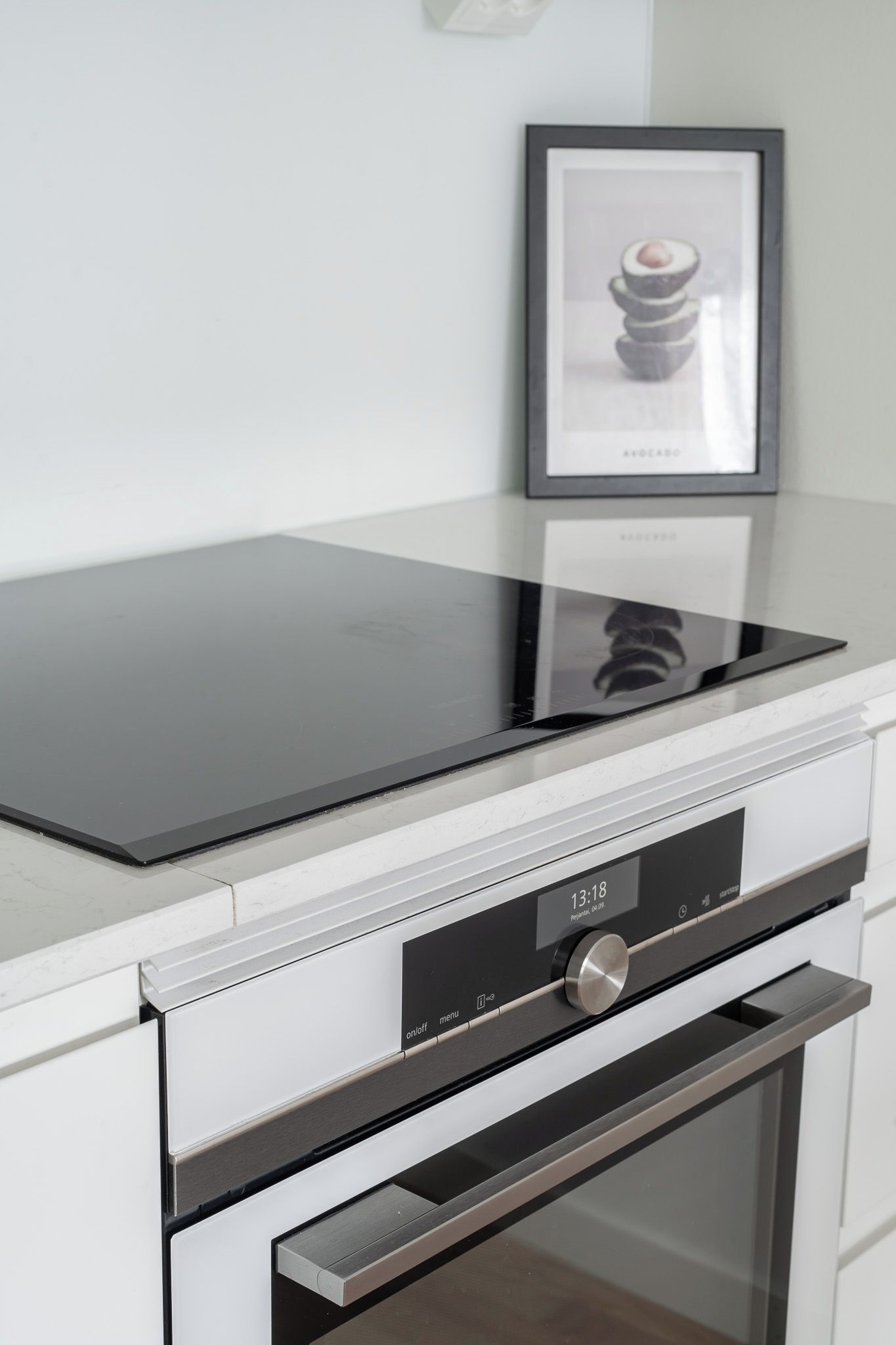 Una cocina con una placa de cocción por inducción negra, un horno de acero inoxidable y un cuadro de un aguacate en la pared.