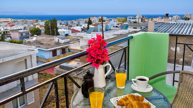 A balcony with a table set for breakfast, including croissants, coffee, and orange juice, overlooking a coastal town. - Room25 (Chania)