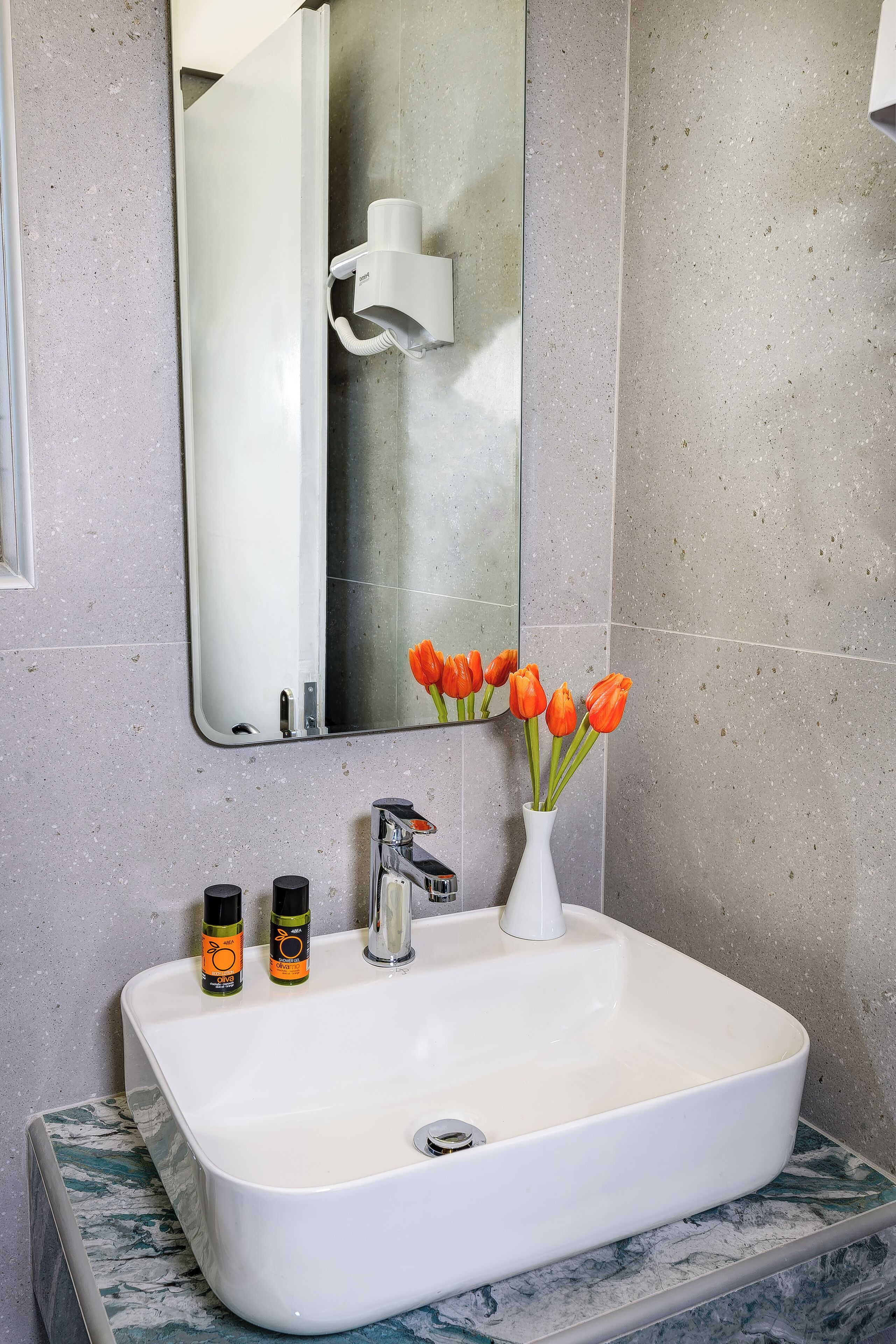 A bathroom with a white sink, a mirror, and a vase with orange tulips. There are two bottles of orange and black soap on the sink.