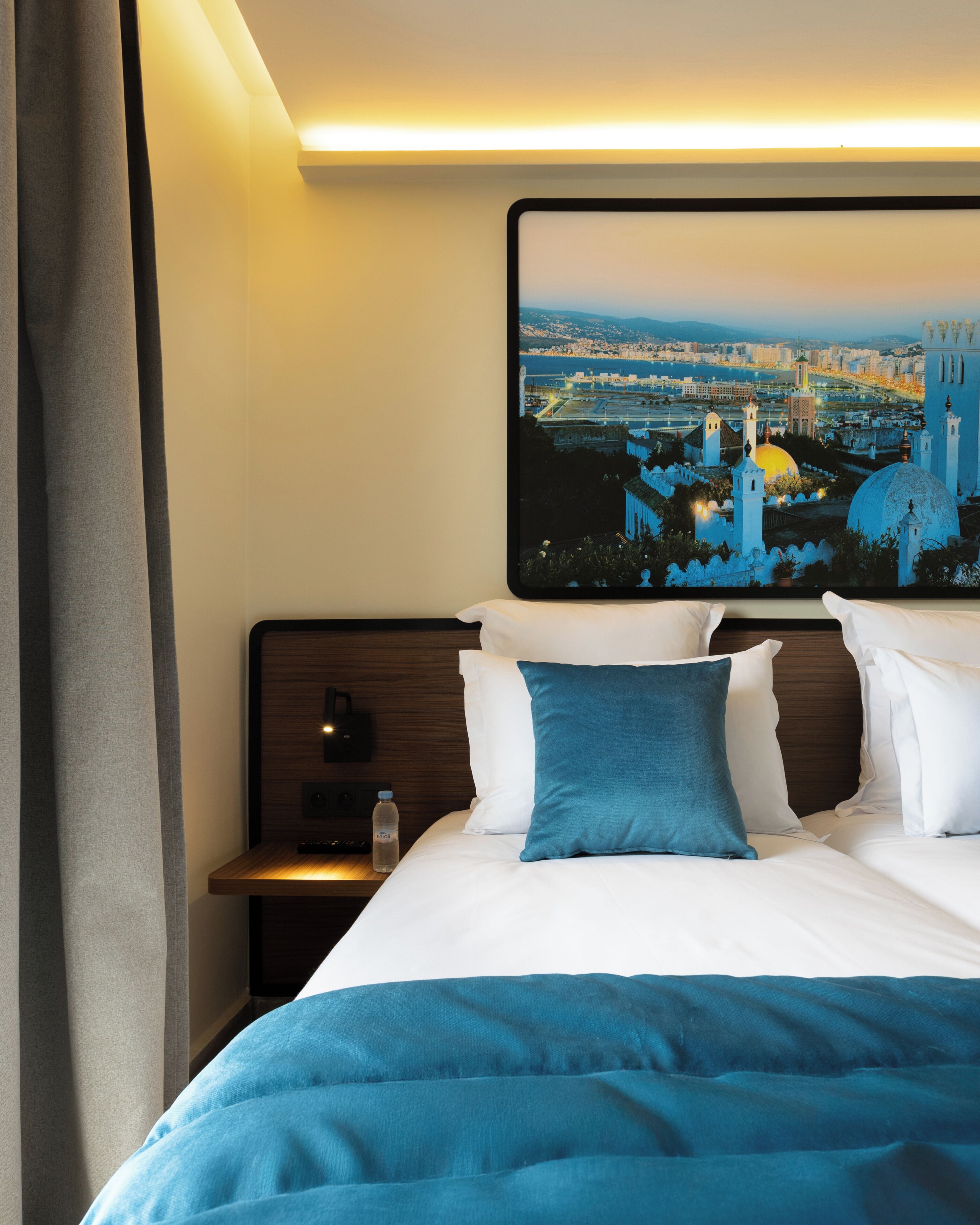 A hotel room with a bed, pillows, a nightstand, a water bottle, and a cityscape painting on the wall.